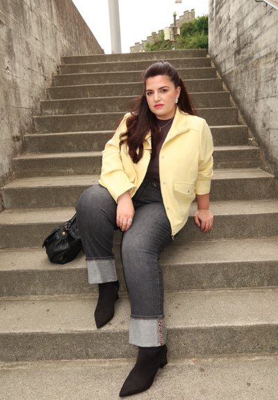 Mujer con cabello oscuro y largo, vestida con una chaqueta amarilla y jeans arremangados, sentada en escaleras de concreto al aire libre al lado de un bolso negro.