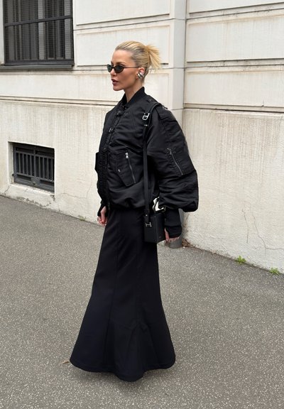 Mujer con gafas de sol y auriculares, lleva una chaqueta acolchada negra, una falda larga negra y lleva un bolso negro mientras camina por una acera urbana.