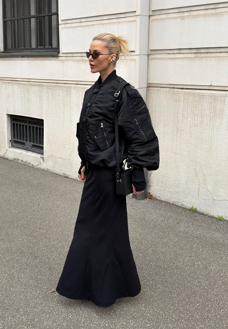 Femme portant des lunettes de soleil et des écouteurs, vêtue d'une doudoune noire, d'une longue jupe noire, et portant un sac à main noir en marchant sur un trottoir urbain.