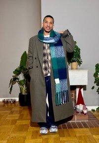 Manteau long vert olive, écharpe à carreaux bleus avec franges, chemise à carreaux, jean bleu et baskets grises. Intérieur avec plantes et décoration.