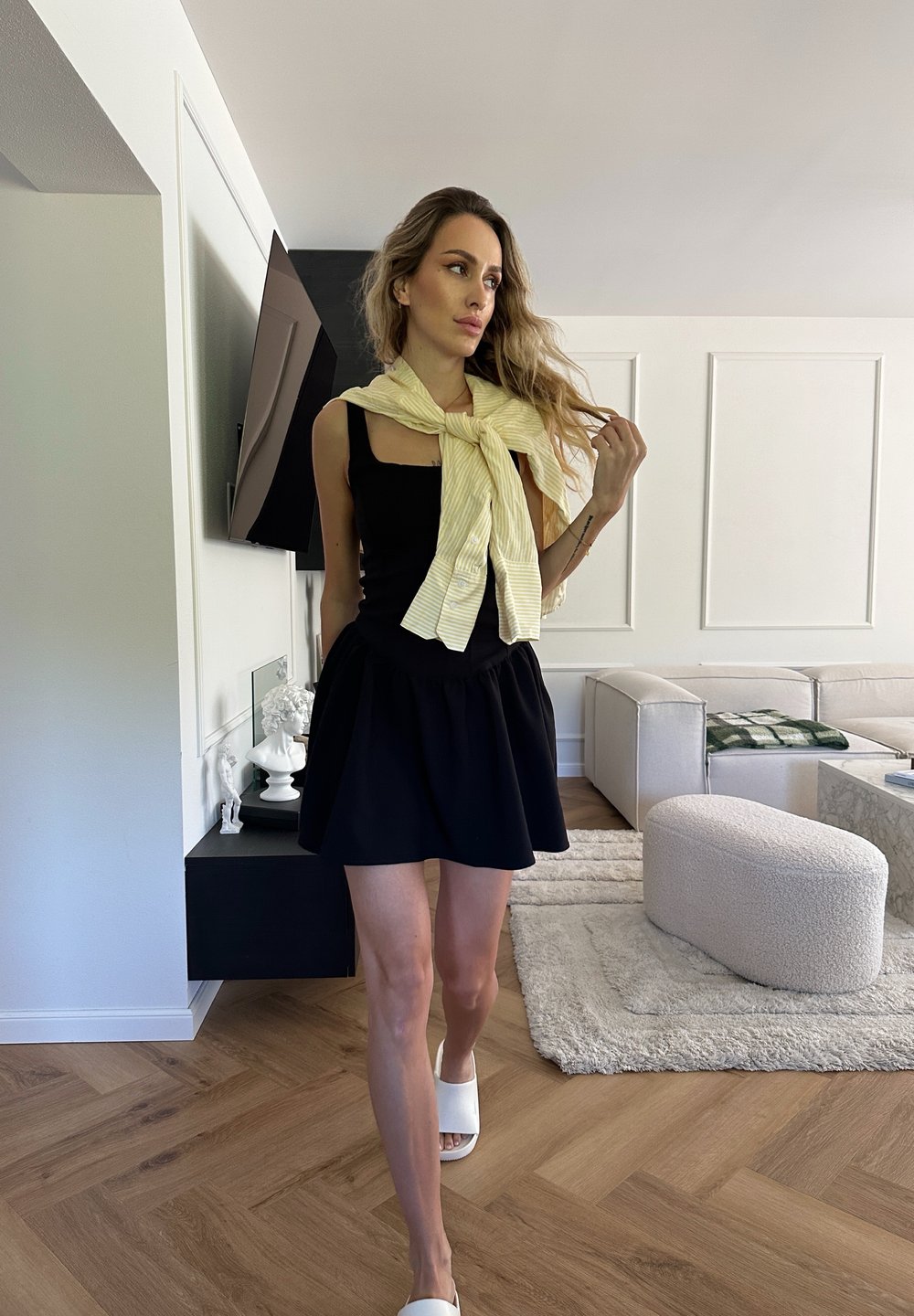 Vestido preto com decote quadrado e saia balonê, combinado com um lenço amarelo listrado. Modelo a usar calçado branco, piso de madeira quente, decoração moderna.