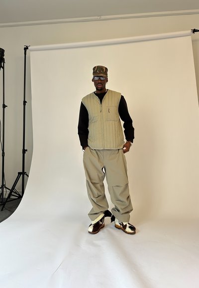 Hombre vestido con un chaleco acolchado beige, camiseta negra de manga larga, pantalones beige holgados, zapatos de cuero de vaca de caña alta y gorra de camuflaje, de pie en un estudio con un fondo blanco.