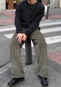 Zwarte cardigan met knopen, olijfgroene corduroy wijde broekspijpen en zwarte schoenen. Zittend op een paal bij een zebrapad met een stoep op de achtergrond.