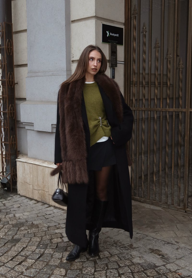 Femme debout sur une rue pavée portant un long manteau noir avec un col en fourrure marron, un pull vert, une jupe noire, des bottes, et tenant un petit sac à main noir.