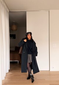 Femme portant un béret noir, un long manteau noir, une jupe grise, des collants noirs et des bottines à talons noires, debout à l'intérieur sur un fond neutre.