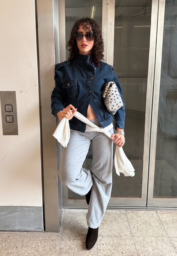 Femme aux cheveux bouclés et grandes lunettes de soleil portant une veste en denim foncé, un pantalon gris, tenant une écharpe à pois et nouant un tissu blanc autour de la taille.