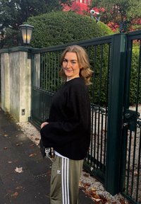 Femme aux cheveux blonds mi-longs portant un pull noir et un pantalon de survêtement Adidas vert, debout près d'une clôture métallique verte en plein air.