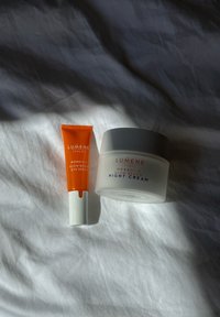 Lumene ihonhoitotuotteet: oranssi silmäseerumi tuubissa ja pyöreä valkoinen yövoidepurkki, jossa on vaaleanharmaa kansi, molemmat ryppyisellä valkoisella kankaalla.
