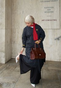 Tunika lunga a righe blu scuro con dettagli dorati, abbinata a pantaloni neri. Tacchi bianchi e una borsa marrone completano il look. Sciarpa rossa annodata al collo.