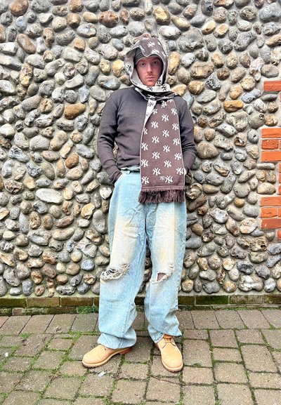 Bufanda marrón con patrón blanco, suéter negro de canalé, jeans claros desgastados y botas marrones. Fondo de pared de piedra con ladrillos.