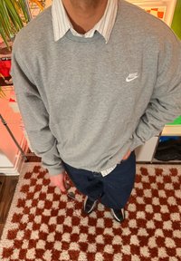 Sweat-shirt gris en coton avec logo Nike blanc, superposé à une chemise à col rayée bleu clair et blanc, associé à un pantalon bleu foncé et des baskets.