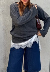 Femme portant un pull gris foncé, une chemise blanche à dentelle, un large jean bleu foncé, et tenant un petit sac à main bordeaux sur un fond gris.