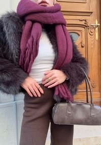 Femme portant un manteau en fourrure foncé, une écharpe épaisse violette, un haut blanc, un pantalon marron, des bagues dorées, tenant un sac à main en cuir marron près d'une porte en bois.