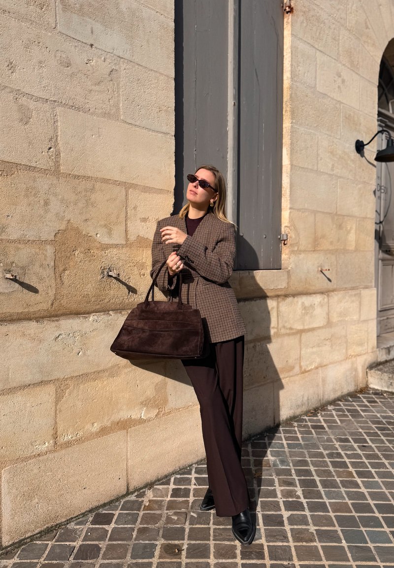 Sacou maro în carouri peste un top închis la culoare, asortat cu pantaloni largi de culoare închisă și pantofi negri cu vârf ascuțit. Ține o geantă maro închis din piele întoarsă.