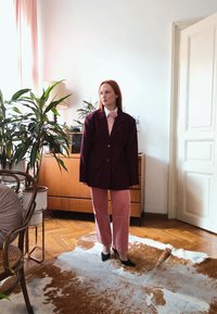 Femme portant un blazer oversize sombre, une blouse rose avec un détail en forme de rose, un pantalon rose et des talons noirs, se tenant dans une pièce baignée de lumière avec des plantes et des meubles en bois.