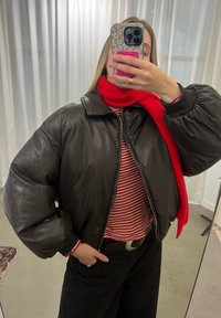 Jaqueta bomber de couro castanho com mangas volumosas, fecho de zíper e punhos pretos. Combinada com um cachecol vermelho de malha e uma blusa listrada por baixo.