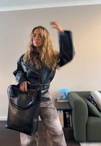 Femme aux longs cheveux bouclés portant une veste en cuir noire, tenant un grand sac noir, debout dans une pièce avec un canapé vert et une lampe bleue.
