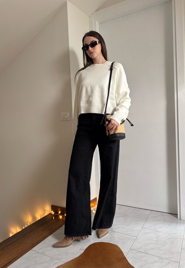 Moteris juodose platėjantose kelnėse, balto megztinio, dėvi smėlio spalvos aukštakulnius, juodus akinius nuo saulės, laikanti mažą pintinę rankinę patalpoje prie uždarytų durų.