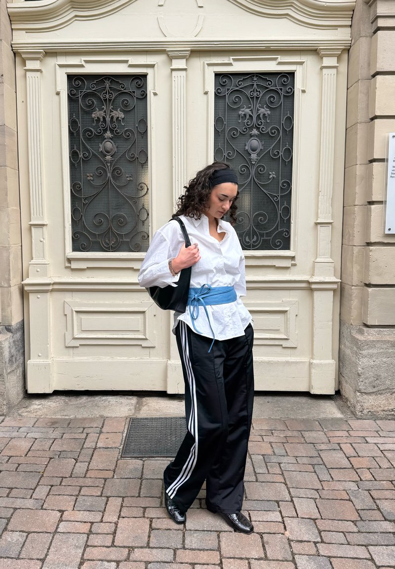 Femme en chemise blanche et pantalon noir à rayures blanches, debout dans une rue pavée devant une porte beige ornée, regardant vers le bas.