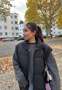 Juodas pūkuotas liemenė virš pilkos megztinio, su širdies pakabučiu. Atsipalaidavęs kirpimas, ilgomis rankovėmis ir kišenėmis.