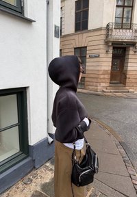 Άτομο που φοράει μαύρο φούτερ με κουκούλα, μπεζ παντελόνι και κρατάει μαύρη τσάντα στέκεται σε καμπυλωτό πεζοδρόμιο κοντά σε μπεζ κτίριο.