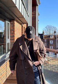 Personne portant un trench-coat marron foncé et une casquette NY noire, tenant une pochette tressée marron, debout sur un balcon avec des immeubles en briques et un ciel dégagé en arrière-plan.