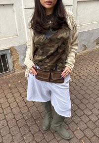 Top camouflage con una stella nera, cardigan beige, pantaloni corti grigi, stivaletti di gomma verde oliva, borsa a cintura in suede marrone con cuciture bianche.