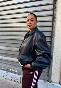 Casaco de couro preto com fecho éclair à frente, fit oversized e mangas compridas. Combinado com calças cor bordeaux com riscas brancas, segurando uma clutch preta trançada.