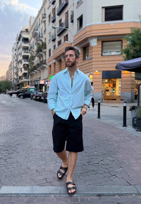 Homme en chemise bleu clair et short noir marchant dans une rue de la ville près des bâtiments au crépuscule.