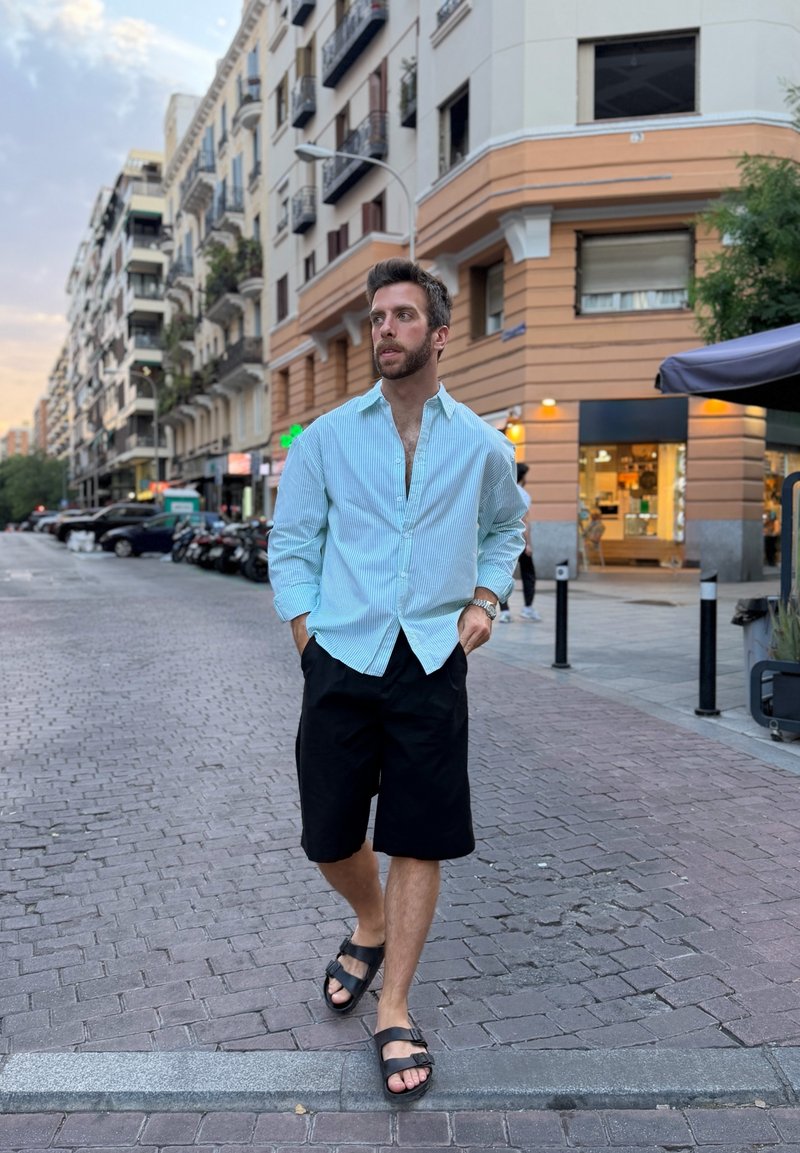 Homme en chemise bleu clair et short noir marchant dans une rue de la ville près des bâtiments au crépuscule.