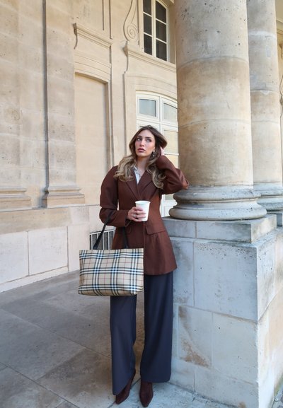 Mujer con blazer marrón y pantalones anchos azul marino, sosteniendo un bolso de cuadros y una taza de café, apoyada en una columna de piedra en un entorno arquitectónico.