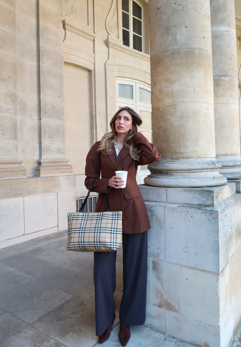 Femme en blazer marron et pantalon large bleu marine, tenant un sac à carreaux et une tasse de café, appuyée contre une colonne en pierre dans un cadre architectural.