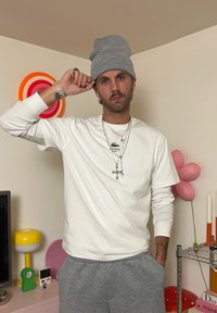 Hombre con una gorra gris, camiseta de manga larga blanca y pantalones de chándal grises, de pie en el interior cerca de una decoración colorida y una estantería de metal.