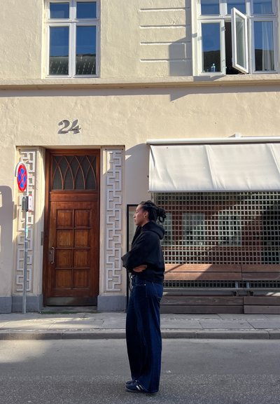Puerta de madera con acentos geométricos, número 24 arriba. Pared beige, gran ventana con un cristal abierto y toldo gris sobre un banco de madera.