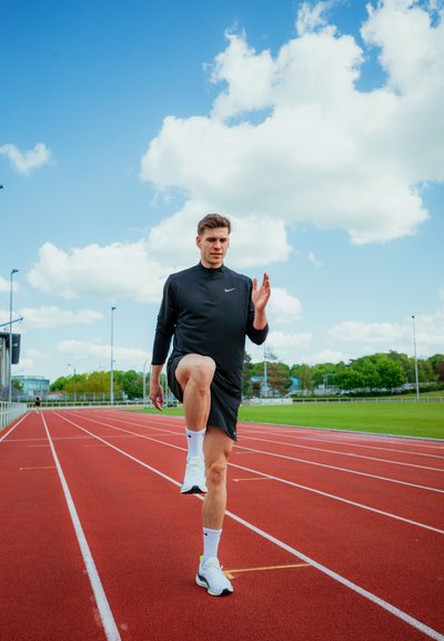 Atleta con camiseta de manga larga negra y pantalones cortos realiza un estiramiento de calentamiento en una pista de atletismo roja, con árboles verdes de fondo.