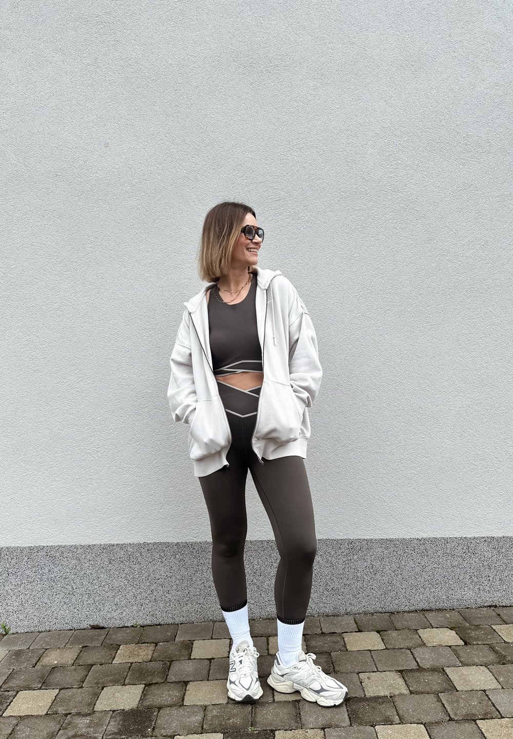 Femme portant des leggings noirs et un crop top avec un sweat à capuche blanc oversize, chaussettes blanches et baskets, debout sur un pavé en pierre près d'un mur gris.