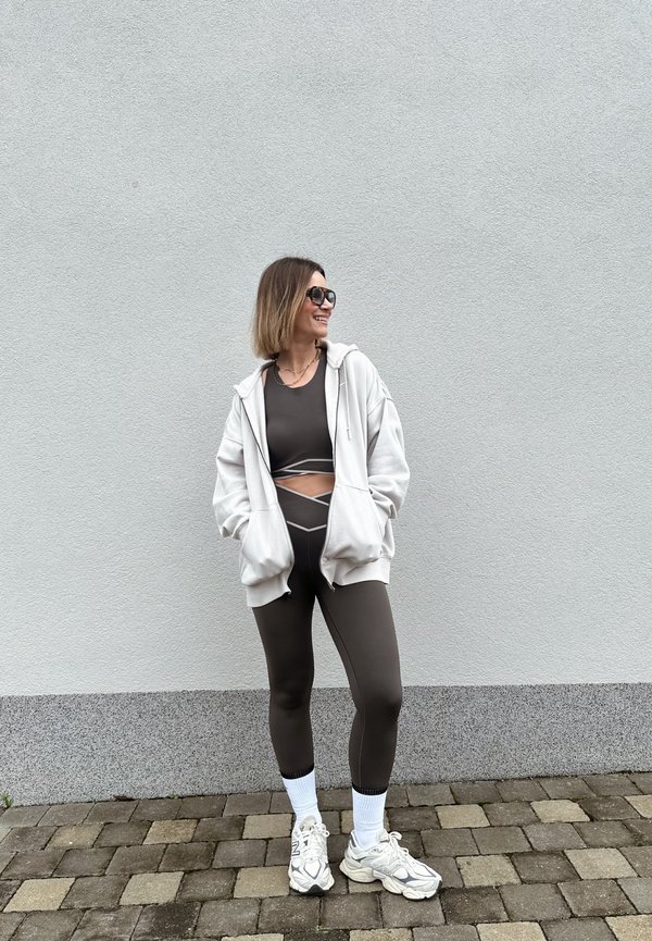 Femme portant des leggings noirs et un crop top avec un sweat à capuche blanc oversize, chaussettes blanches et baskets, debout sur un pavé en pierre près d'un mur gris.