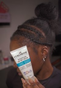 Person med flettet hår og knute holder et rør med Curlsmith Weightless Air Dry Cream nær ansiktet, med produktetiketten tydelig synlig.