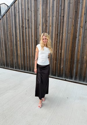 Mujer rubia con tatuajes que lleva una blusa blanca, falda negra y tacones blancos, de pie sonriendo junto a una pared de tablones de madera verticales.