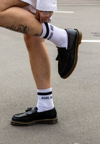 Scarpe in pelle nera con nappine, cuciture gialle, abbinate a calzini bianchi con strisce nere e testo. Lo sfondo è un pavimento grigio.