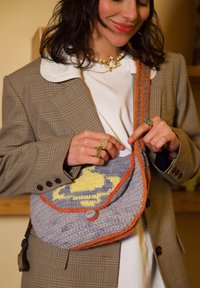 Femme portant un blazer à carreaux et une chemise blanche, tenant un sac violet crocheté avec un motif jaune et une bordure orange sur l'épaule.