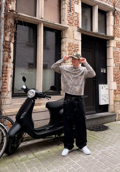 Joven ajustándose una gorra de camuflaje, de pie junto a un scooter negro en una calle empedrada con un edificio de ladrillo y una puerta detrás de él.