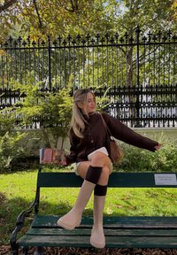 Rudas megztas kardiganas, kremiškos šortai, smėlio spalvos platformos botai, rudi kelių ilgio kojinės, laiko knygą, sėdi ant žalios suolelio su augalijos fonu.