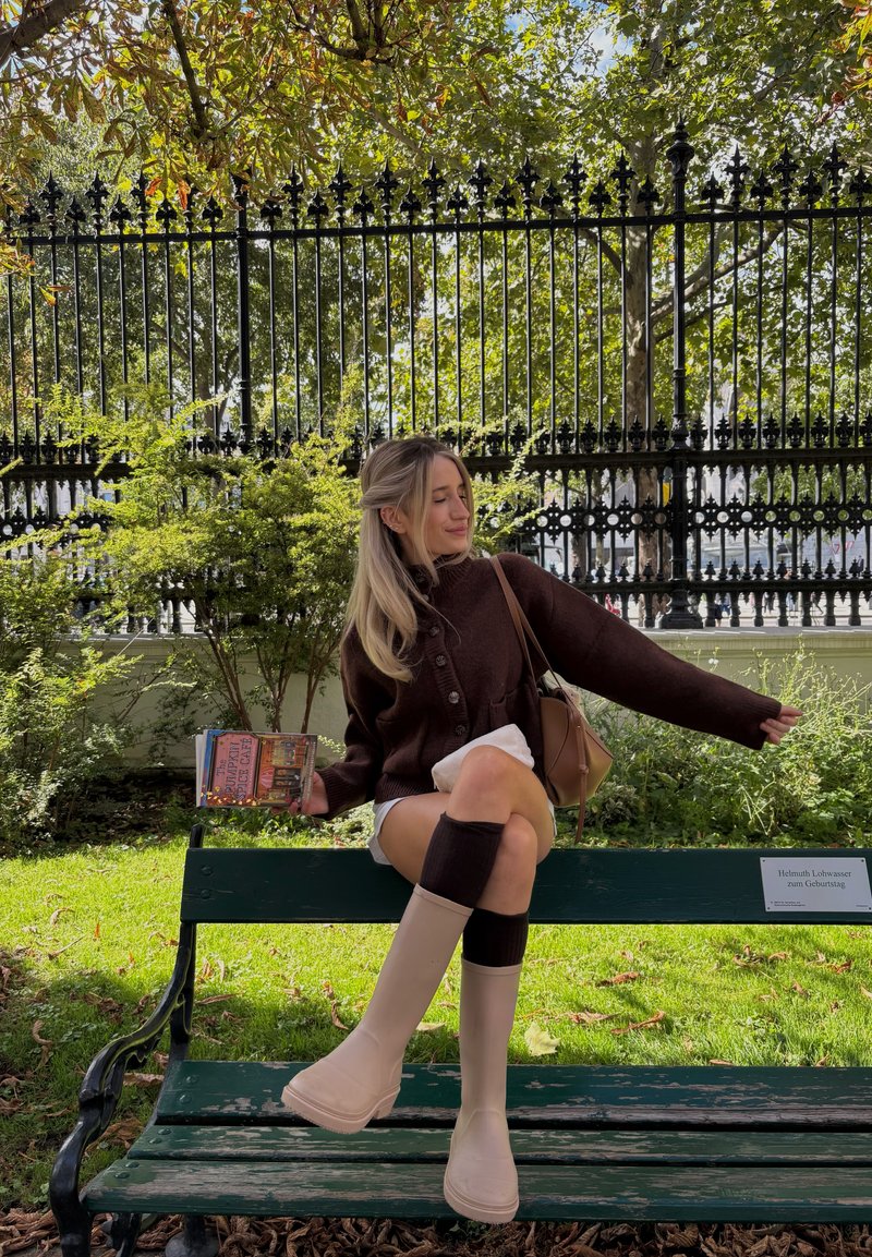 Brun strikket cardigan, cremefarvede shorts, beige platform støvler, brune knæ-længde sokker, holder en bog, siddende på en grøn bænk med baggrund af løv.