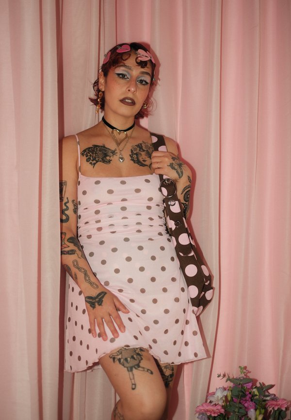 Jeune personne tatouée porte une robe rose à pois et un sac assorti, debout devant des rideaux roses avec une petite composition florale à proximité.