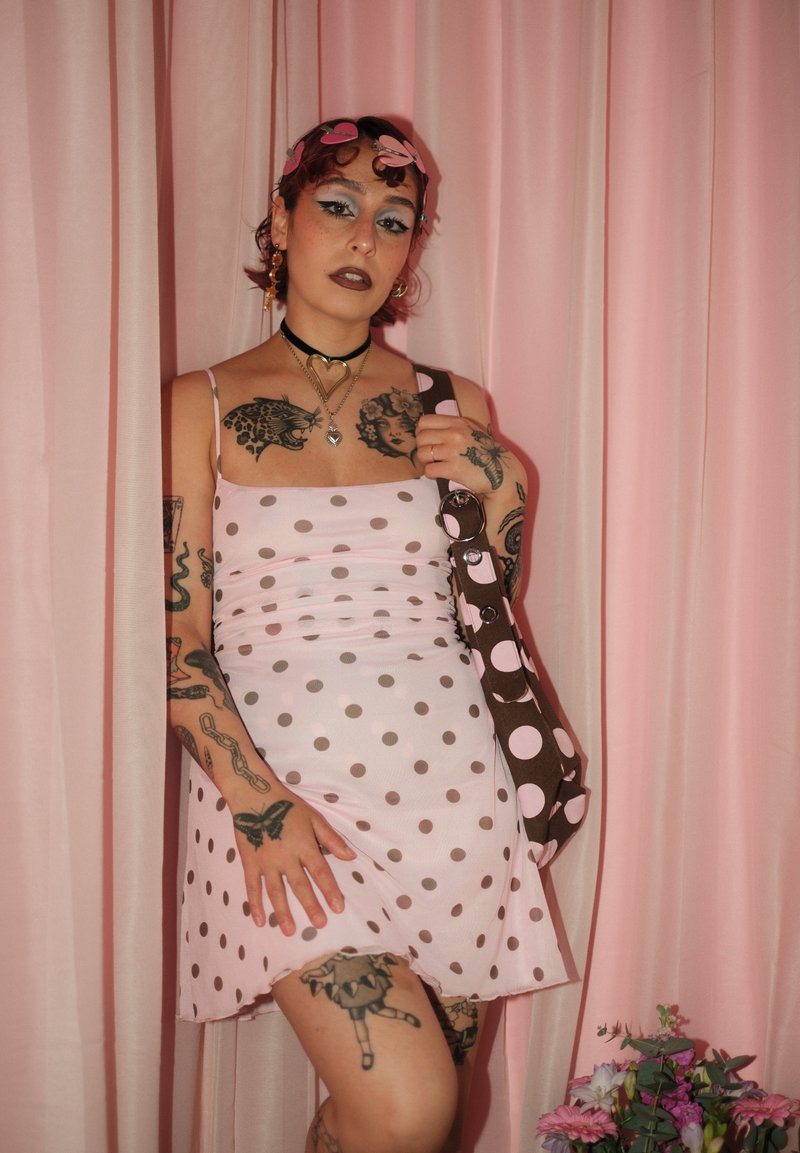 Jeune personne tatouée porte une robe rose à pois et un sac assorti, debout devant des rideaux roses avec une petite composition florale à proximité.