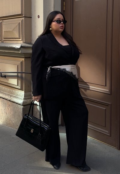 Mujer con traje negro y cinturón de pañuelo beige, gafas de sol negras, pendientes dorados y bolso negro junto a una puerta marrón al aire libre.