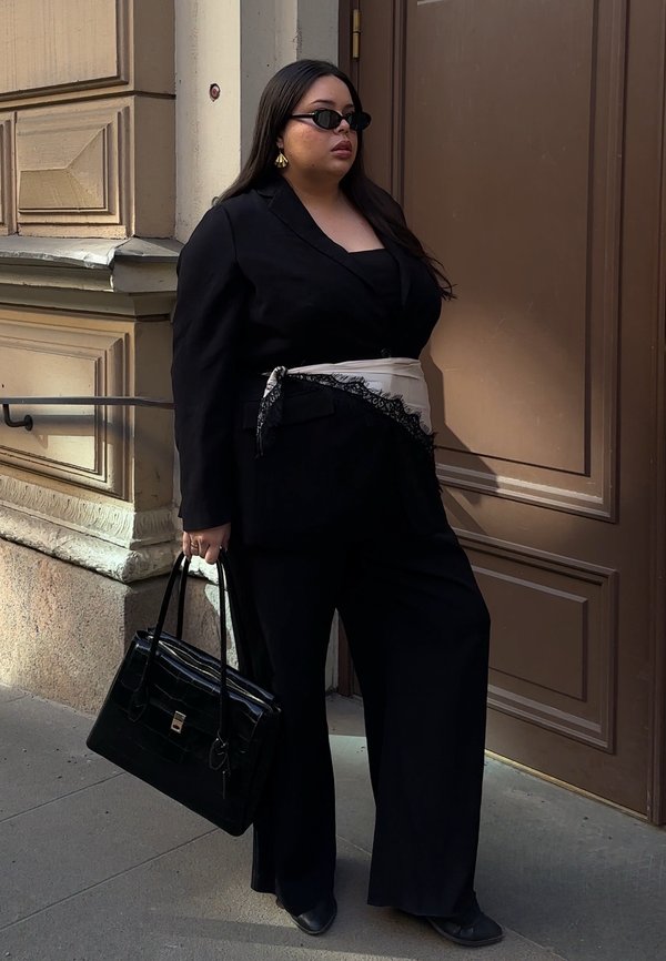 Femme en costume noir avec ceinture foulard beige, lunettes de soleil noires, boucles d'oreilles en or et sac à main noir, debout près d'une porte marron à l'extérieur.