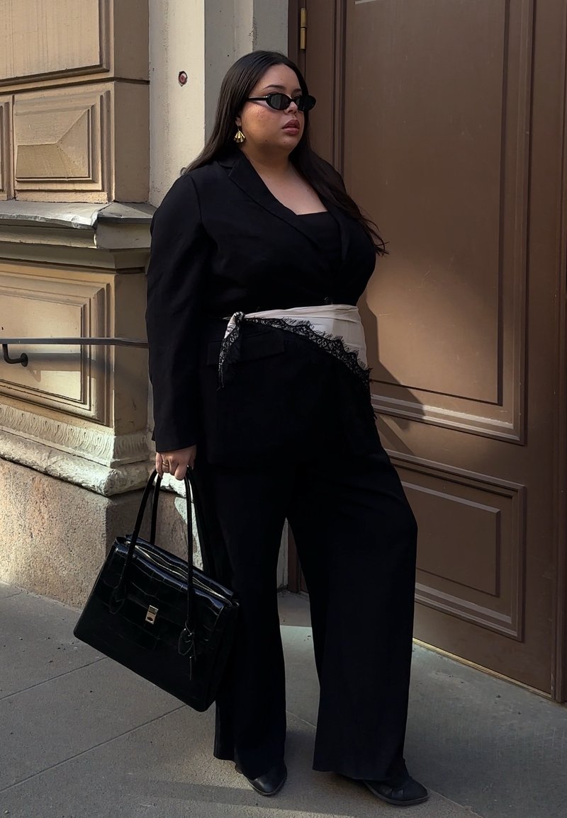Femme en costume noir avec ceinture foulard beige, lunettes de soleil noires, boucles d'oreilles en or et sac à main noir, debout près d'une porte marron à l'extérieur.