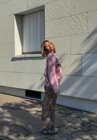 Suéter rosa claro de gran tamaño con texto negro, pantalones de pierna ancha con estampado de leopardo y sandalias grises. El fondo presenta una pared blanca texturizada.